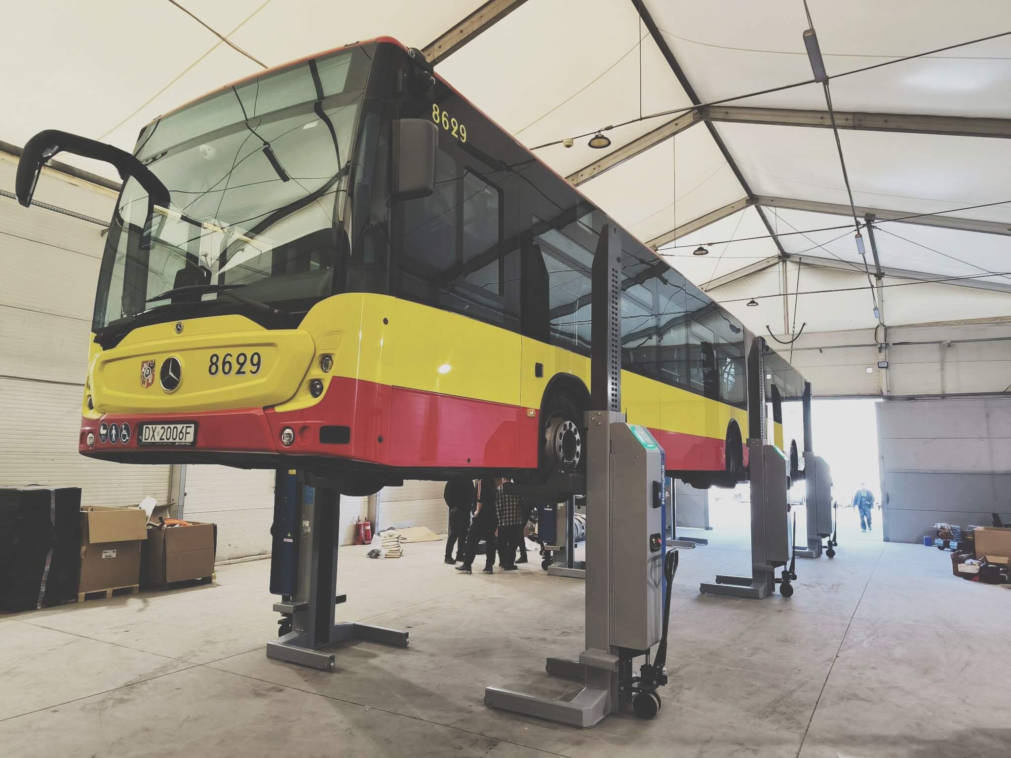 Realizacja Anwa-Tech: Autobus przegubowy Mercedes-Benz uniesiony na profesjonalnych podnośnikach kolumnowych w hali serwisowej. Widoczny przód pojazdu i precyzyjne rozmieszczenie kolumn dźwigowych